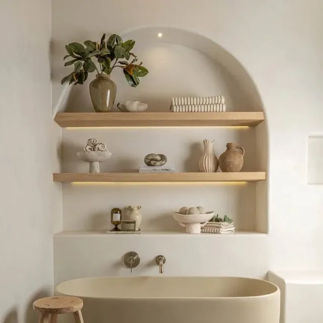 An arched niche in pale tadelakt over a freestanding bathtub, its softly lit wooden shelves holding vases, a plant, and folded towels, a wooden stool beside the tub