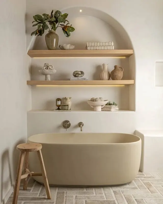 An arched niche in pale tadelakt over a freestanding bathtub, its softly lit wooden shelves holding vases, a plant, and folded towels, a wooden stool beside the tub