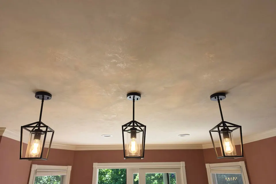 A kitchen ceiling in softly polished bone Marmorino, its cloudy movement catching the light above three lantern pendants and terracotta walls, with trees in the windows below