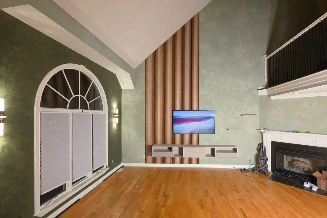 A two-story living room in gray-green limewash, with oak panels behind the TV, floating shelves and cabinets, and a darker green clay wall around an arched window