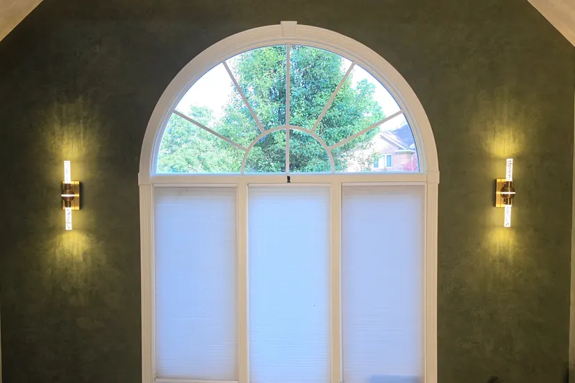 The inset wall in smoky green Roman clay around a tall arched window, a sconce glowing on each side, in a Geist entry