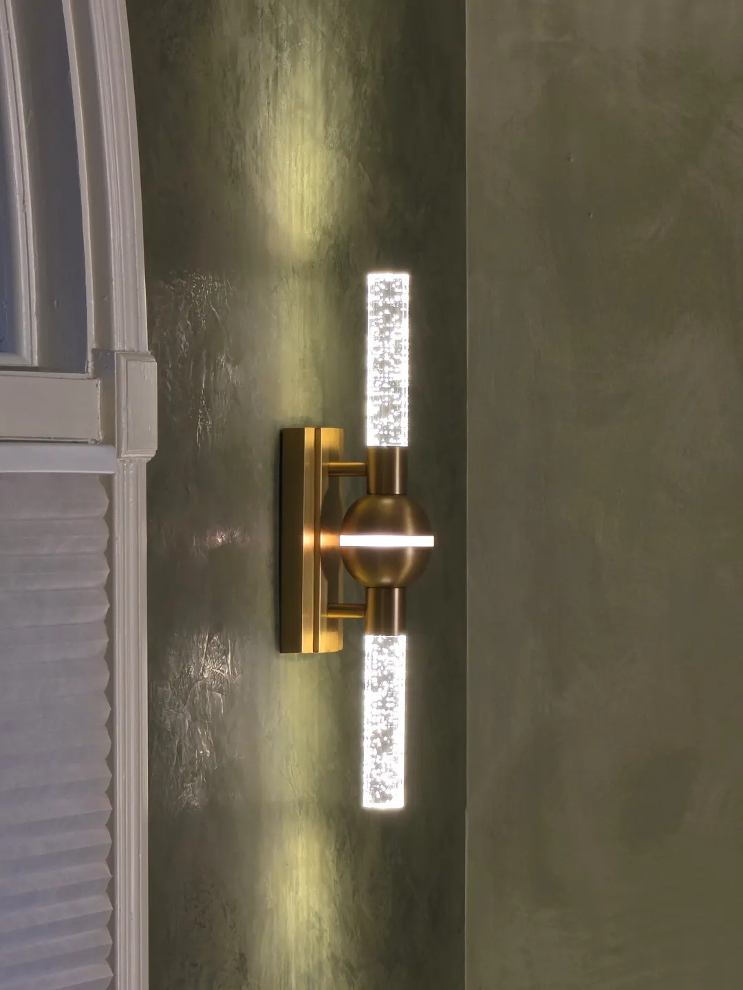 A crystal and brass sconce glowing on smoky green Roman clay beside an arched window, in a Geist entry