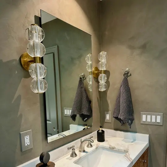 Bathroom walls in a dark olive limewash around a mirror, glass and brass sconces, and a marble-topped vanity, in a home