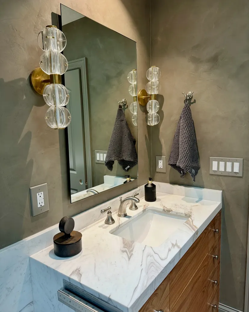 Bathroom walls in a dark olive limewash around a mirror, glass and brass sconces, and a marble-topped vanity, in a home