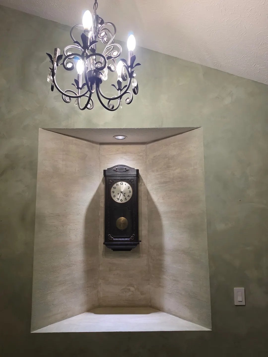 A lit alcove lined in faux travertine with a wall clock inside, set into a soft green wall under a chandelier, in a hall