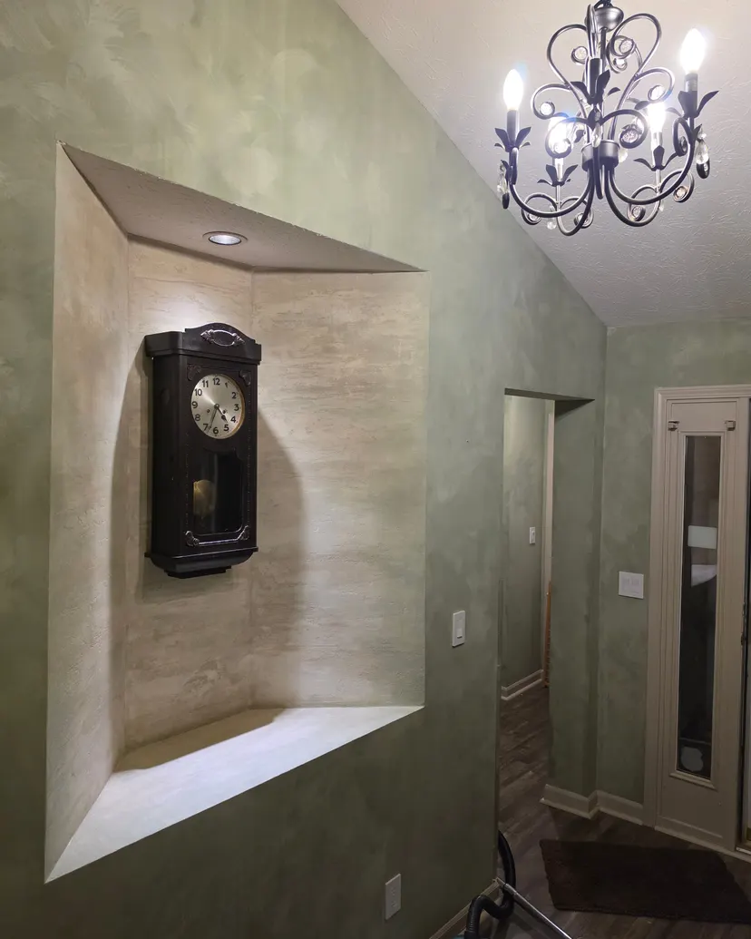 The lit faux travertine alcove with its wall clock, seen from the hall under a black scrolled chandelier, the walls a soft, mottled green