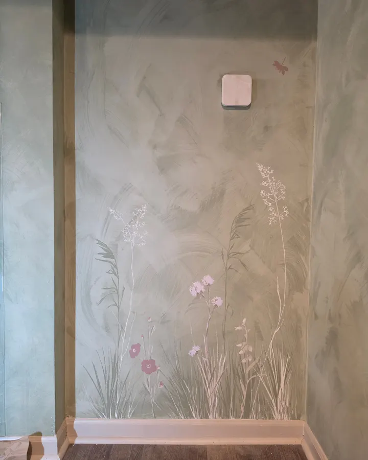 Wildflowers and grasses in pink, white, and green painted up from the baseboard inside a soft green alcove, a small pink dragonfly above them
