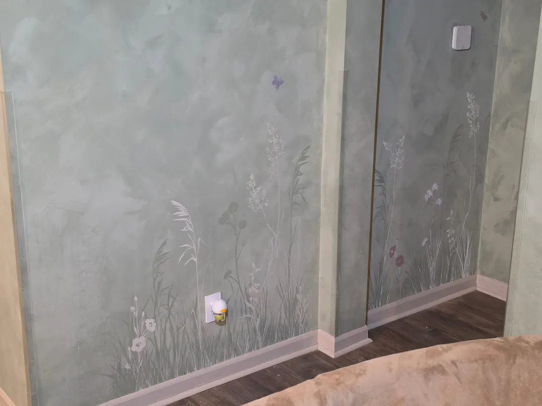 Wildflowers and tall grasses painted up from the baseboards on a soft green limewash wall, a small butterfly above them, in a home