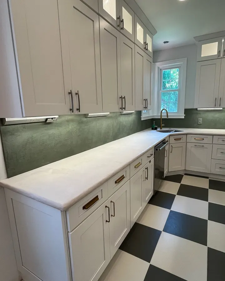 A soft green microcement backsplash under white upper cabinets, the outlets tucked beneath the cabinets, above a pale microcement counter