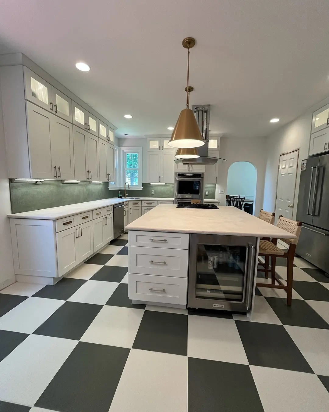 Pale microcement countertops on an island and along the walls, a soft green microcement backsplash, brass pendants, and a black and white checkered floor, in a kitchen