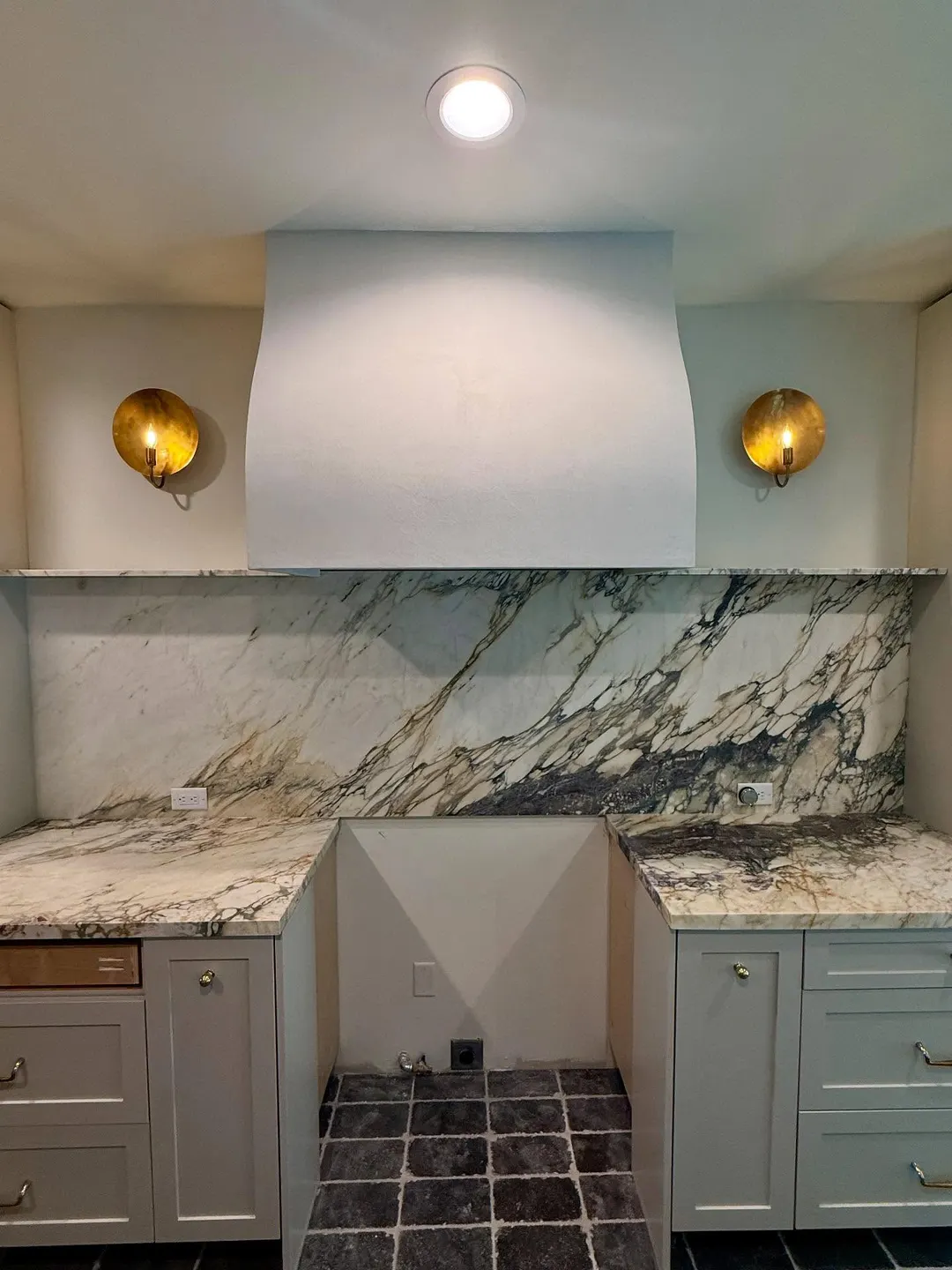 A pale, curved plaster range hood between two brass sconces, above a boldly veined backsplash, the range not yet in, in a kitchen