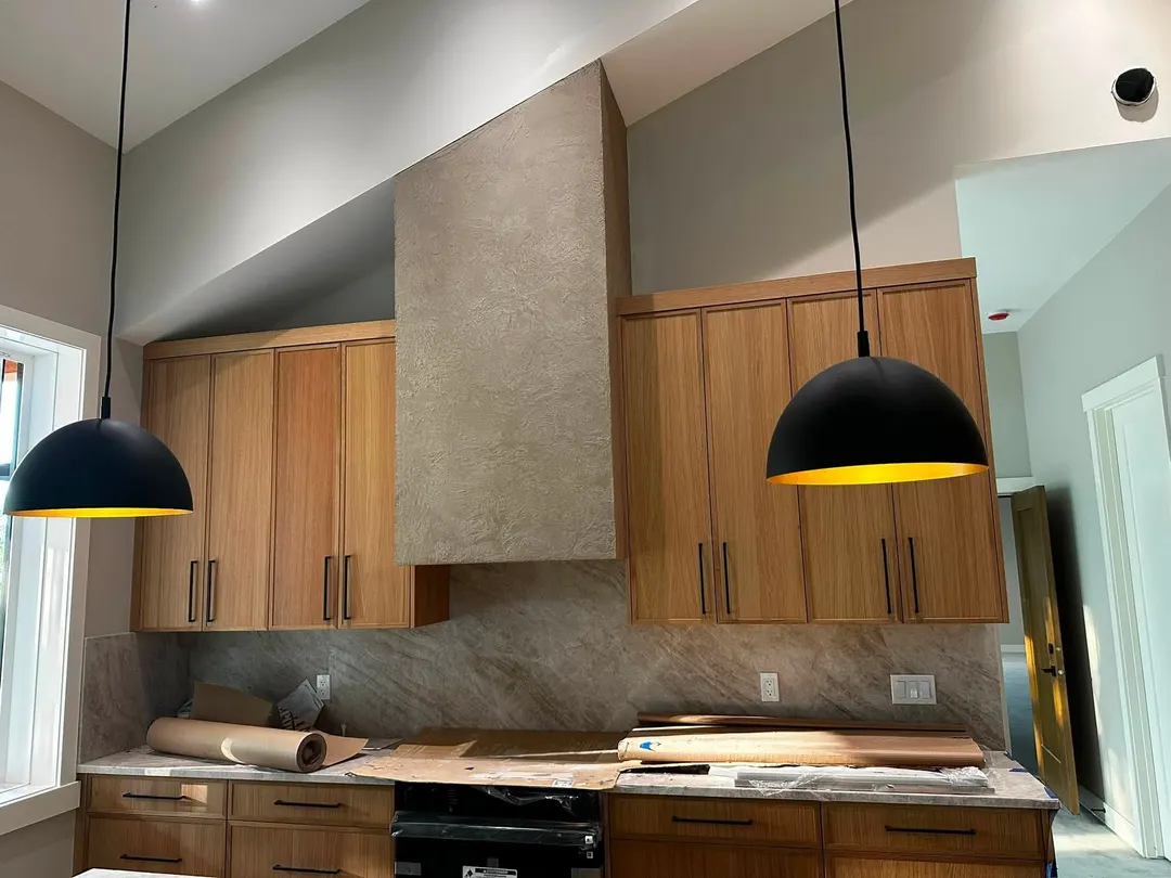 A tall gray plaster range hood with a stone-like texture rising toward a vaulted ceiling, between warm wood cabinets and two black pendants, in a kitchen