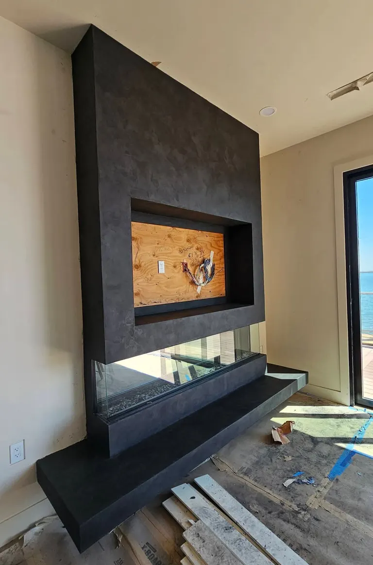A charcoal plaster fireplace surround with a recessed niche above a glass-sided fireplace and a long floating hearth, new flooring stacked in front, in a home
