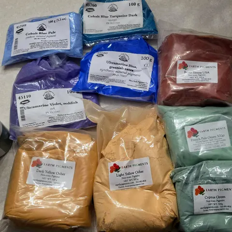 Bags of raw pigment in blues, violets, ochres, reds, and greens on a studio table