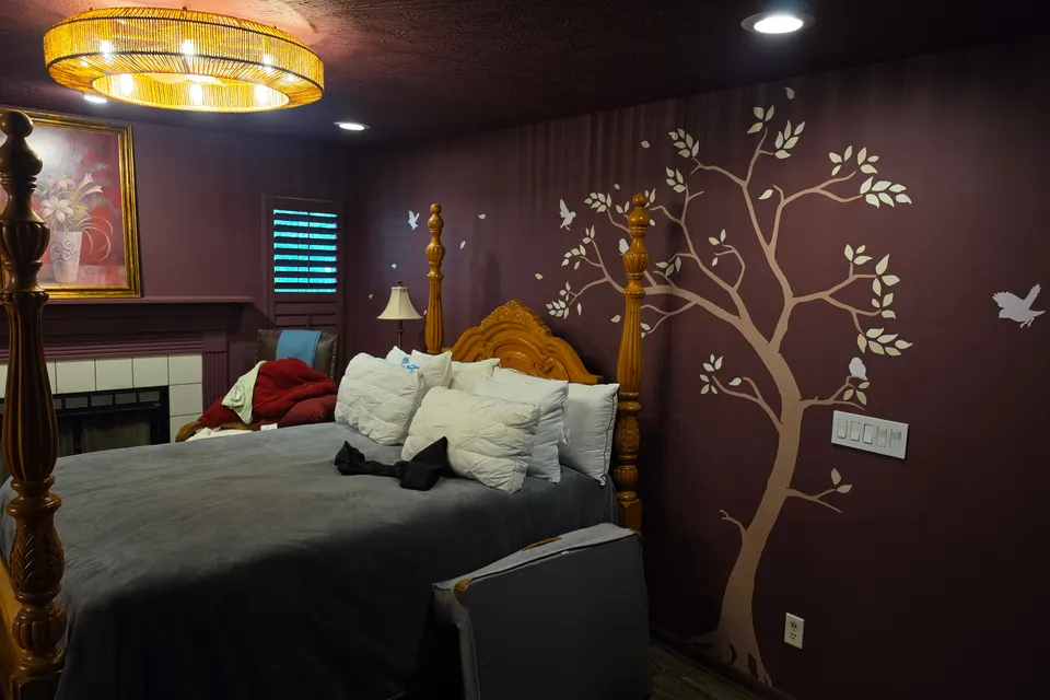 A bedroom in deep oxblood under a glowing woven ceiling light, with a carved four-poster bed and a pale tree painted on the wall, in a Zionsville home