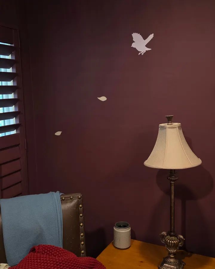 A pale painted bird and two falling leaves on a deep oxblood bedroom wall, above a table lamp, in a Zionsville home