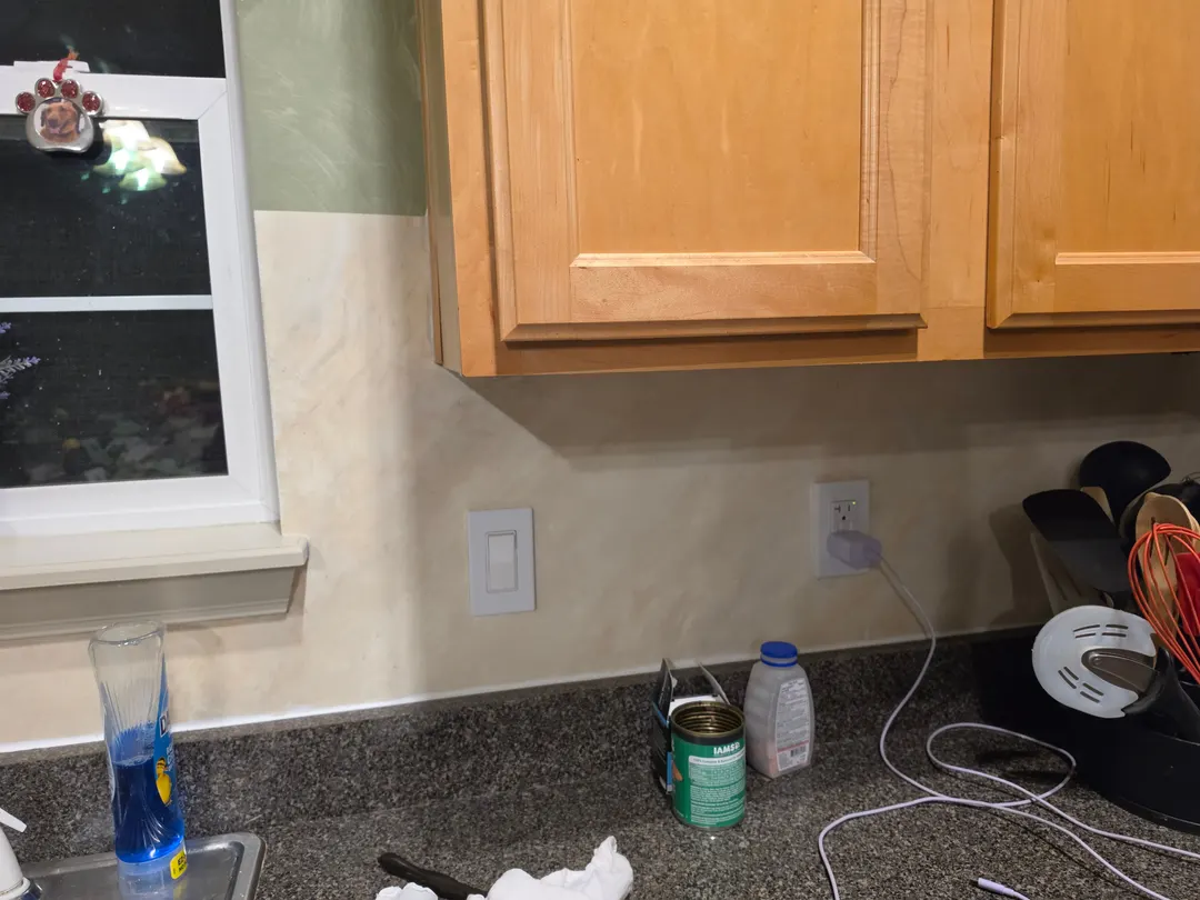 The polished cream marble backsplash under honey-toned wood cabinets beside a window, above a dark speckled counter